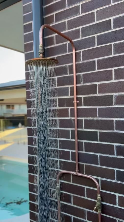 Exposed copper outdoor shower running against a dark brick wall beside a pool